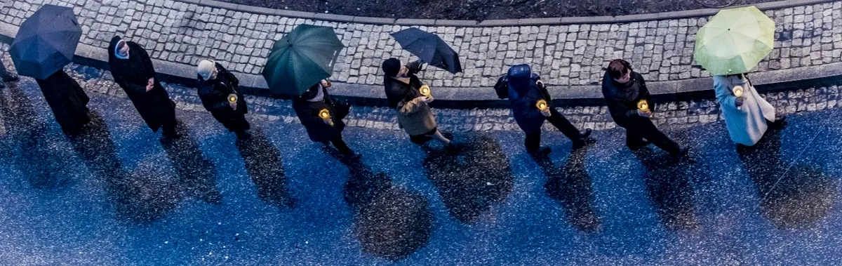 Line of pedestrians with umbrellas and candlelight reflected on wet blue pavement